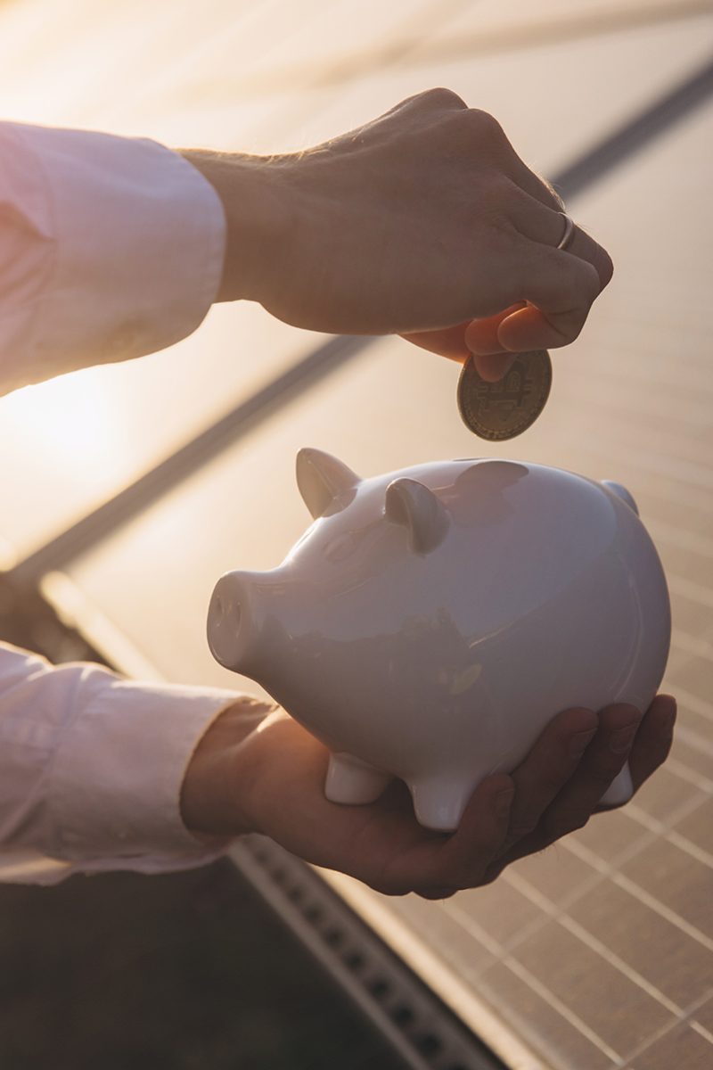A person places a coin into a piggy bank against the backdrop of solar panels at sunset, symbolizing financial savings and renewable energy investments.